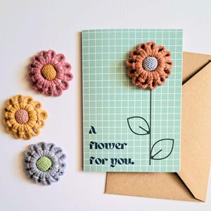 May include: A green card with a grid pattern and a black line drawing of a flower with the text "A flower for you." Three crocheted flowers in pink, yellow, and grey are placed on a white surface.