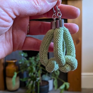 May include: A pair of light green, knotted rope earrings. The earrings have silver-colored metal hooks and cylindrical accents. The knotted design creates a unique, textured appearance. The earrings are held up by a hand.