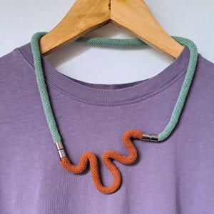 Peut inclure: Un collier crocheté vert et orange avec un design ondulé. Le collier est sur un t-shirt violet.