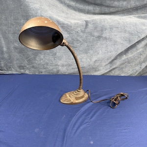 Könnte beinhalten: Eine metallisch braune Schreibtischlampe mit einem kuppelförmigen Lampenschirm, einem flexiblen Schwanenhalsarm und einem stabilen Sockel. Die Lampe verfügt über ein braunes Stromkabel mit Stecker. Die Außenseite des Schirms weist Gebrauchsspuren auf.