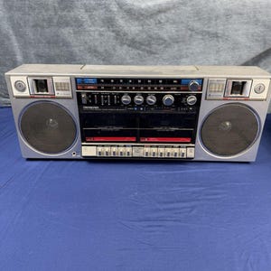 Może przedstawiać: Srebrny przenośny odtwarzacz kasetowy stereo z dwoma kieszeniami na kasety, dwoma okrągłymi głośnikami i wyświetlaczem radiowym. Przedni panel zawiera liczne pokrętła, szereg przycisków oraz nadrukowaną na biało nazwę marki Fisher.