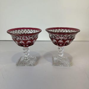 Puede incluir: Dos cuencos de postre de cristal a juego con un borde rojo arándano intenso y un cuenco transparente con patrón de corte de diamante. Cada cuenco se asienta sobre un tallo transparente facetado y una base cuadrada con un diseño de rayos.