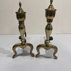 Puede incluir: Un par de morillos de chimenea de latón pulido con una columna central alta y delgada rematada con un remate, apoyada sobre una base curva de tres patas. Cada morillo tiene una barra horizontal de hierro negro que sobresale por detrás.