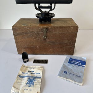 May include: A black Berger dumpy level surveying instrument mounted on a rectangular wooden storage box. Included are a small black metal lens hood, a metal identification plate, a parts list document, and a manual titled Level and Transit-Level.
