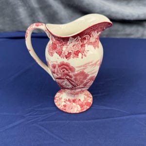 May include: A cream-colored ceramic pitcher featuring a red transferware pattern. The design depicts a landscape with trees, buildings, and floral motifs. The pitcher has a curved handle, a flared rim, and a pedestal base, all decorated with the red print.