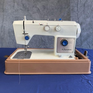 May include: A white Signature sewing machine with a tan base. The machine features a silver control panel with a blue dial labelled Stitch Length Control, two white adjustment knobs on the top, a needle, and a metal throat plate with markings.