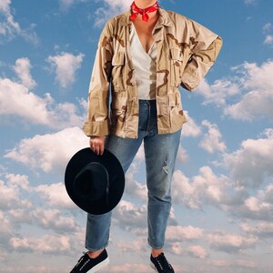 May include: A person wearing a camouflage jacket with a red bandana, blue jeans, and a black hat. The person is standing in front of a blue sky with white clouds.