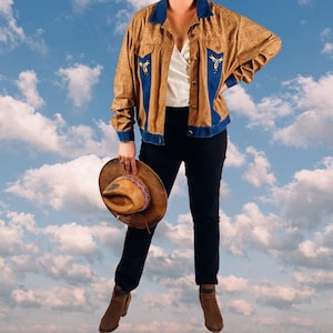 May include: A brown suede jacket with blue denim accents and gold embroidery. The jacket has a western style and features a pointed collar, long sleeves, and a button closure. The model is wearing the jacket with black jeans and a brown cowboy hat.