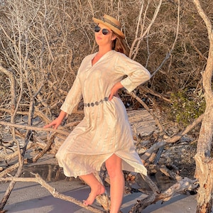 May include: A woman wearing a white dress with a silver belt and a straw hat stands on a beach with driftwood in the background.