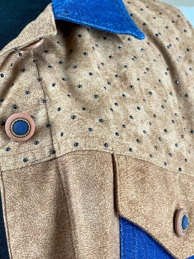 May include: Close-up of a brown suede jacket with black polka dots and a blue denim collar. The jacket has two large buttons on the front, one of which is visible in the image.