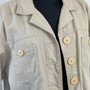 May include: A beige utility jacket with four large, light brown buttons. The jacket has a single chest pocket and two flap pockets.