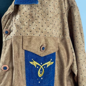 May include: A brown suede jacket with a blue denim collar and a blue denim pocket with gold embroidery. The jacket has a pattern of small black dots.