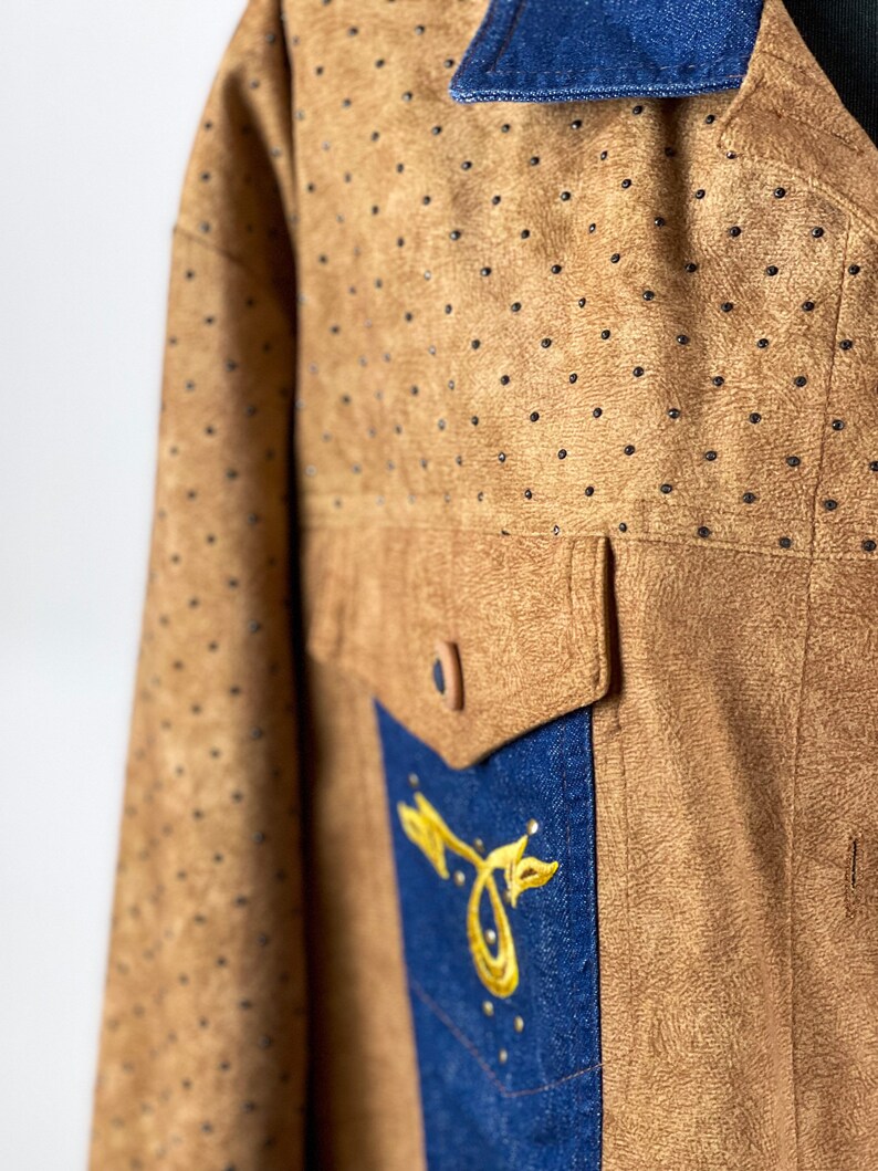 May include: A brown suede jacket with a blue denim collar and a blue denim panel on the front. The jacket has a gold embroidered design on the denim panel.