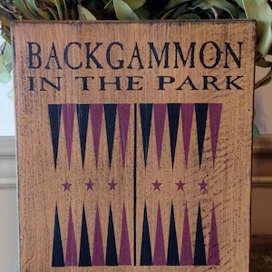 May include: Wooden backgammon game board sign with the text "BACKGAMMON IN THE PARK". The board features black and purple triangles and stars. The sign also includes the text "EVERY FRIDAY 4 O'CLOCK PM HUDSON & SONS GAME BOARDS".