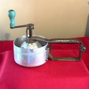 May include: Vintage metal food mill with a perforated bowl and a crank handle. The handle has a light blue wooden grip. The mill is designed for processing fruits and vegetables. The mill is sitting on a red surface.