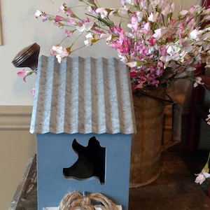 May include: A decorative blue birdhouse with a corrugated metal roof and a pig-shaped opening. The birdhouse has a white base with a rope and wood accent. Pink and white flowers are in a metal bucket in the background.