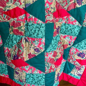May include: A patchwork quilt with a vibrant mix of floral and paisley patterns in shades of red, teal, and beige. The quilt features geometric shapes and a solid red border, creating a cozy and colorful textile.