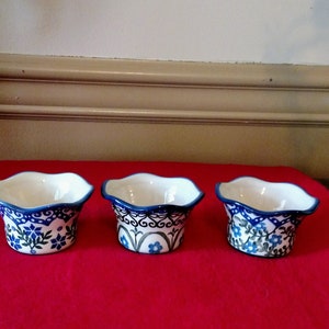 May include: Three small, white ceramic bowls with a blue floral design and scalloped edges. The bowls are arranged on a red surface, showcasing their intricate patterns and vibrant colors. Each bowl has a unique floral pattern.