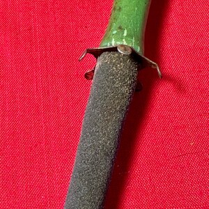 May include: A sharpening tool featuring a textured, dark gray, tapered abrasive rod attached to a smooth, bright green handle. A small, metallic, four-pronged collar connects the handle to the rod, showing signs of wear and minor surface oxidation.