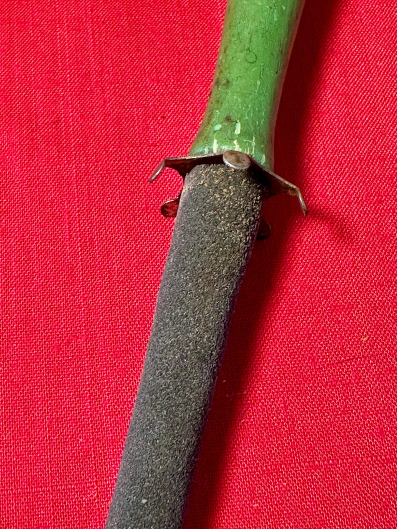 May include: A sharpening tool featuring a textured, dark gray, tapered abrasive rod attached to a smooth, bright green handle. A small, metallic, four-pronged collar connects the handle to the rod, showing signs of wear and minor surface oxidation.