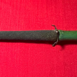 May include: A sharpening tool featuring a dark gray, textured, conical abrasive rod attached to a smooth, green-painted handle. A small, silver-colored metal guard separates the abrasive sharpening section from the handle of the tool.