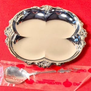May include: A silver-colored, four-sectioned serving dish with ornate edges. A silver spoon is in a plastic wrapper with the text "DEEPSILVER INTERNATIONAL". The dish is on a red surface.