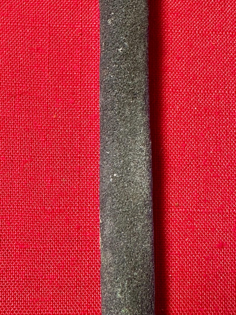 May include: A long, narrow, dark gray strip with a textured, granular surface. The strip is positioned vertically against a bright red background that features a fine, woven, cross-hatched fabric pattern throughout the entire visible area.