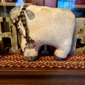 May include: A stuffed sheep figurine with a cream-colored body, gray accents, and a black and brown plaid scarf. The sheep has a decorative berry stem attached to its neck. The figurine is displayed on a red and brown braided rug.