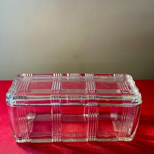 May include: A rectangular, clear glass container with a lid. The glass has a grid-like pattern on the lid and vertical lines on the sides. The container sits on a red surface.