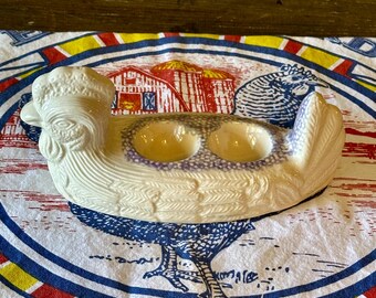 May include: A white ceramic chicken-shaped egg holder with two egg-shaped indentations. The holder has a textured surface and is resting on a white cloth with a poultry-themed design in red, blue, and yellow.