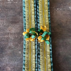 May include: Two rectangular, decorative fabric pieces with vertical stripes in gold and teal. Each piece is adorned with a small bow made of gold and green ribbons. The items are displayed on a dark wooden surface.