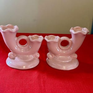 May include: Two matching pink ceramic vases. Each vase has a curved horn shape with a ruffled top and a circular opening in the middle. The base is rounded with small decorative spheres. The vases are displayed on a red surface.