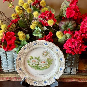 May include: A decorative plate with a floral design, featuring white water lilies and green leaves. The plate has a white background with a blue and beige border. The text "Compliments of Your Grocer, A. E. Seckinger." is printed on the plate. The plate is displayed on a black stand.