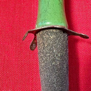 May include: A tool featuring a smooth, bright green handle connected to a dark, textured, cylindrical abrasive sharpening stone. A thin, weathered metal guard with curved edges separates the green handle from the rough, gray stone surface.