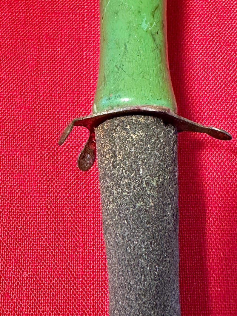May include: A tool featuring a smooth, bright green handle connected to a dark, textured, cylindrical abrasive sharpening stone. A thin, weathered metal guard with curved edges separates the green handle from the rough, gray stone surface.