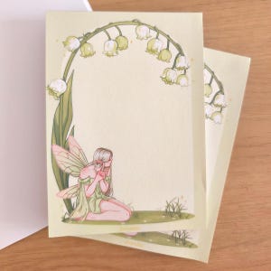 Puede incluir: Una hoja rectangular de papel de carta amarillo pálido con la ilustración de un hada de alas translúcidas sentada sobre una zona de hierba verde. Un arco de flores blancas de lirio de los valles y tallos verdes enmarca la parte superior.