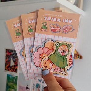 Może przedstawiać: Zestaw trzech naklejek z motywem Shiba Inu. Naklejki przedstawiają Shiba Inu w zielonym stroju żaby, Shiba Inu w r&oacute;żowym stroju kwiatu i Shiba Inu w misce z ramenem. Naklejki znajdują się na brzoskwiniowym tle z wzorem siatki.