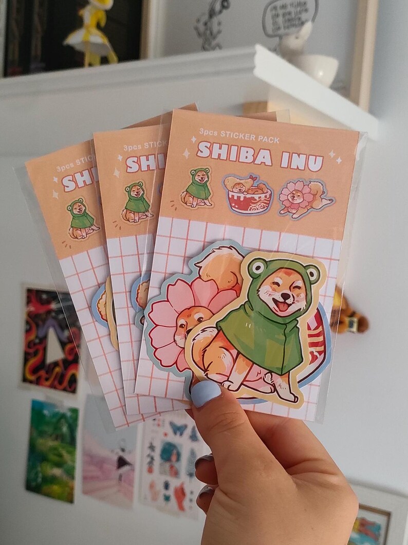 Może przedstawiać: Zestaw trzech naklejek z motywem Shiba Inu. Naklejki przedstawiają Shiba Inu w zielonym stroju żaby, Shiba Inu w r&oacute;żowym stroju kwiatu i Shiba Inu w misce z ramenem. Naklejki znajdują się na brzoskwiniowym tle z wzorem siatki.