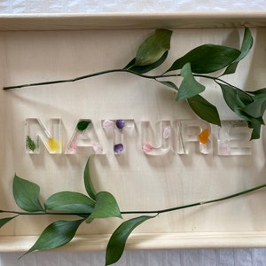 May include: A wooden tray with the word "NATURE" spelled out in clear resin letters. Each letter has pressed flowers and leaves inside. The tray is decorated with sprigs of green leaves.
