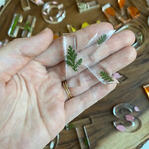 May include: A clear resin letter K with green pressed leaves inside. The letter is being held in a hand.