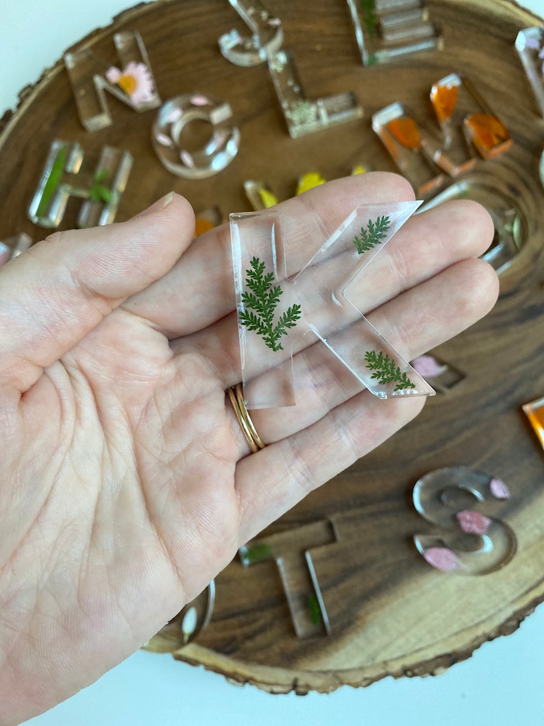 May include: A clear resin letter K with green pressed leaves inside. The letter is being held in a hand.