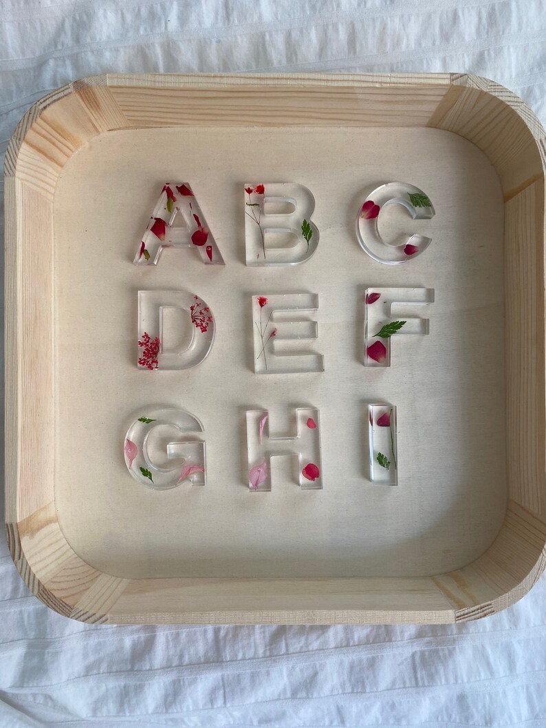 May include: Nine clear resin letters, A through I, with pressed flowers and petals inside. The letters are arranged in three rows on a wooden tray.