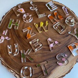 May include: A set of clear resin alphabet letter tiles with pressed flowers and leaves inside. The letters are arranged on a wooden surface.