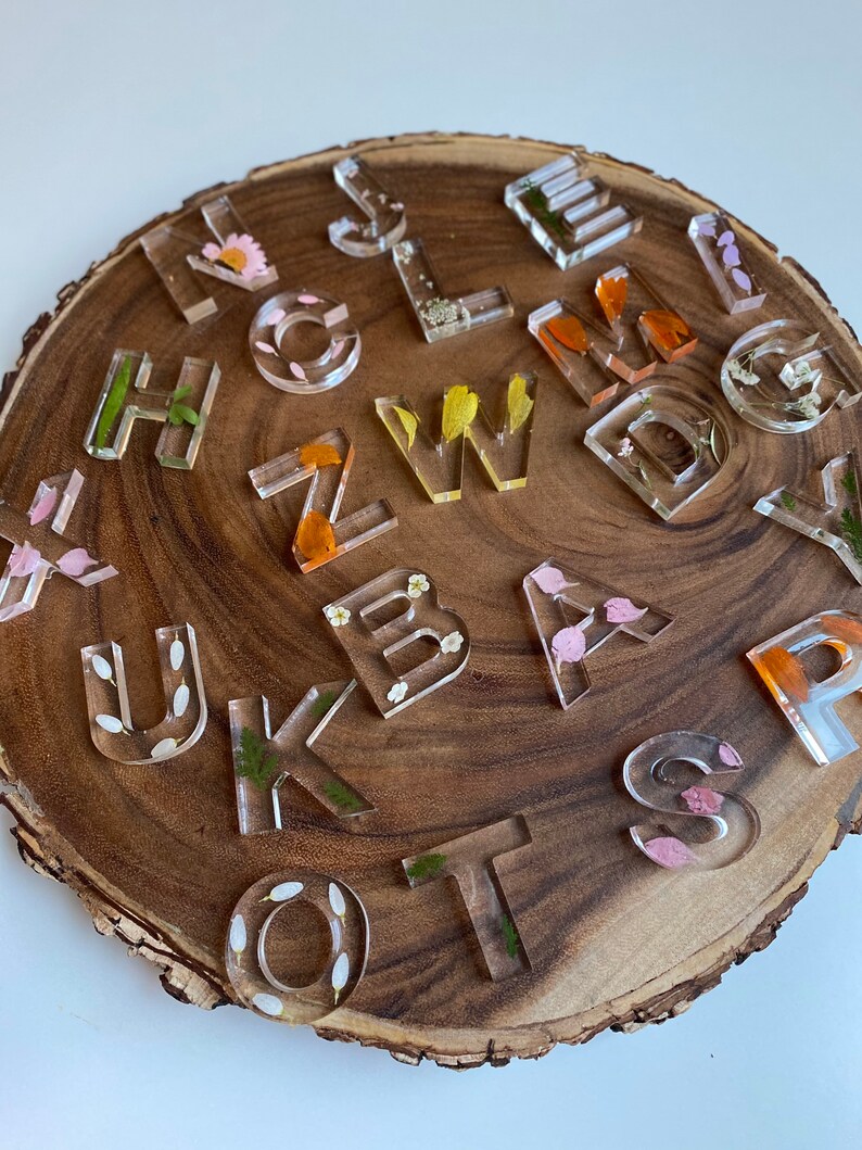 May include: A set of clear resin alphabet letter tiles with pressed flowers and leaves inside. The letters are arranged on a wooden surface.