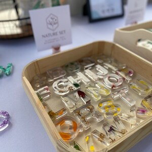 May include: A wooden box filled with clear resin alphabet letter charms. Each letter has pressed flowers and leaves inside. The box is labeled "Nature Set".