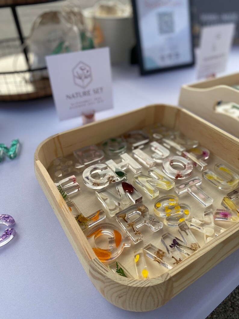 May include: A wooden box filled with clear resin alphabet letter charms. Each letter has pressed flowers and leaves inside. The box is labeled "Nature Set".