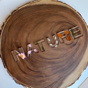 May include: A wooden round slice with the word "NATURE" spelled out in clear resin letters. The letters are filled with pressed flowers and leaves.