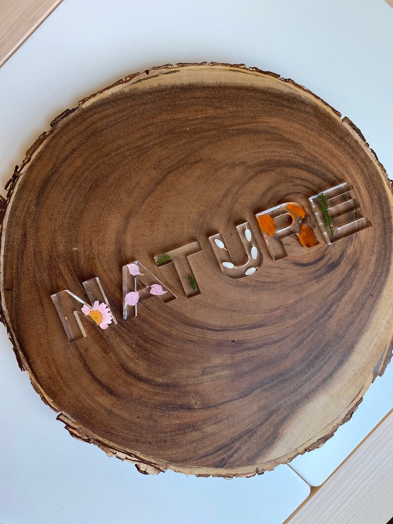 May include: A wooden round slice with the word "NATURE" spelled out in clear resin letters. The letters are filled with pressed flowers and leaves.