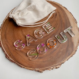 May include: Clear resin letters spelling "SAGE SCOUT" with pressed flowers inside. The letters are on a wooden slice with a white drawstring bag.