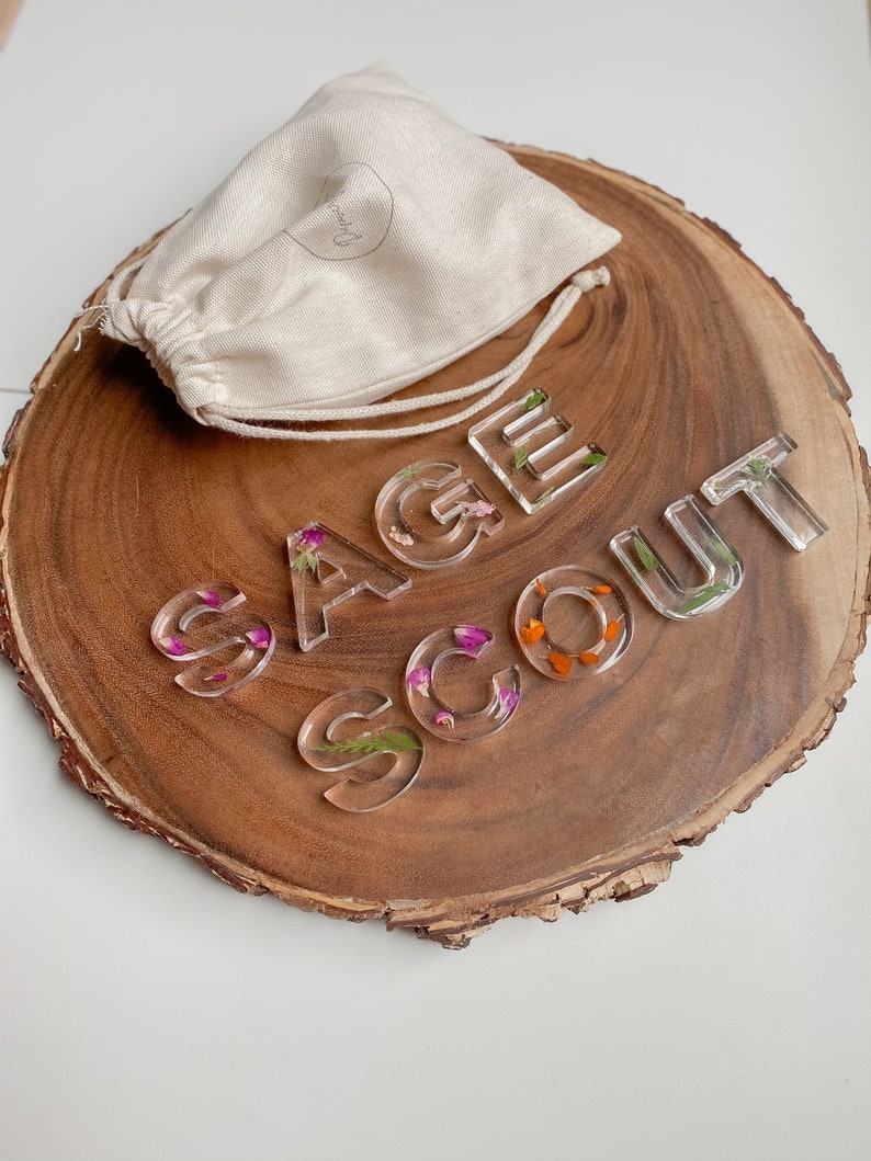 May include: Clear resin letters spelling "SAGE SCOUT" with pressed flowers inside. The letters are on a wooden slice with a white drawstring bag.